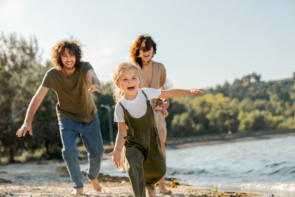 Happy young family having fun on the beach. Little cute girl running along a beach being chased by her parents. - 写真・画像
