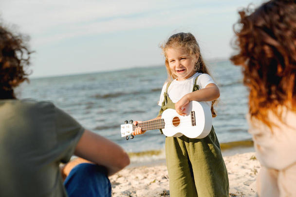 Young man and woman sitting on a beach while their cute little daughter playing on ukulele for them - Photo, Image