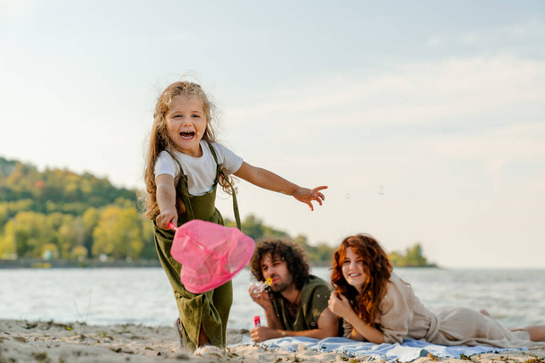 Portrait of happy little girl playing with butterfly net with parents having fun on backgroung on the beach - Φωτογραφία, εικόνα