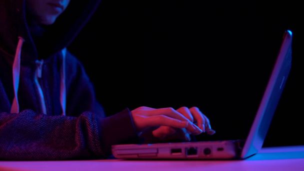 Young woman in a hood with a laptop. Hacker makes a hack through a laptop. Blue and red light falls on a woman on a black background. - Foto, afbeelding
