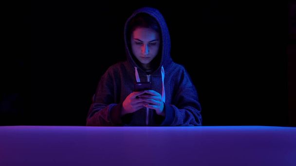 Young woman in a hood with a smartphone. Hacker makes a hack through the phone. Blue and red light falls on a woman on a black background. - Fotoğraf, Görsel