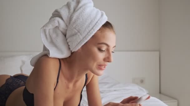 Pleased pretty brunette woman in lingerie using smartphone and enjoying the moment while lying on bed at home - Materiaali, video