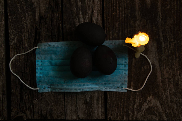 máscara médica y velas de llama de huevo negro de color en el día de Pascua en una vieja mesa de madera durante la pandemia
 - Foto, Imagen
