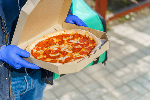 Пицца с салями и сыром в картонной коробке. Безопасность доставки в синих медицинских перчатках из ресторана. Карантин для остановки коронавируса ковид 19
 - Фото, изображение