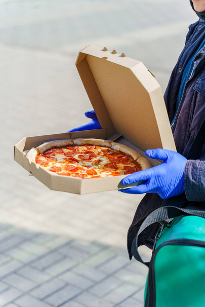 Pizza with salami and cheese in cardboard box. Safety delivery in blue medical gloves from restaurant. Quarantine for stop coronavirus covid 19 - Photo, Image