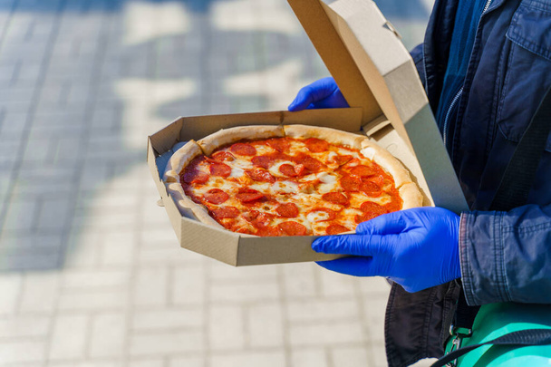 Biztonságos pizzafutár az étteremből. Élelmiszer futár orvosi kesztyűben. Elszigetelt fotó pizzáról sajttal és szalámival. Állítsa le a pandémiás coronavirus covid 19 - Fotó, kép