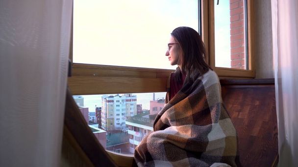 Egy fiatal nő ül az ablaknál. Egy nő ül egy ablakpárkányon, kockás ruhában, és kinézett az ablakon. Az ablak előtt egy fényes nap.. - Fotó, kép