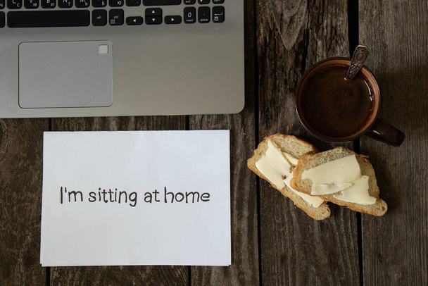 koffie en wit brood en boter op een houten tafel en de woorden in het Engels zitten thuis op wit papier en een laptop - Foto, afbeelding