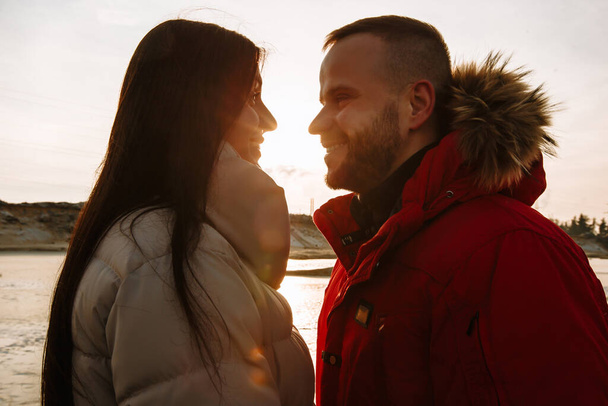 Young people in down jackets on a frozen lake at sunset. A man looks into a girl's face with love - Foto, imagen