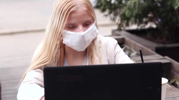 A young girl sits on a street in a cafe with a protective mask of white color. Girl looking at a laptop. Video - Footage, Video