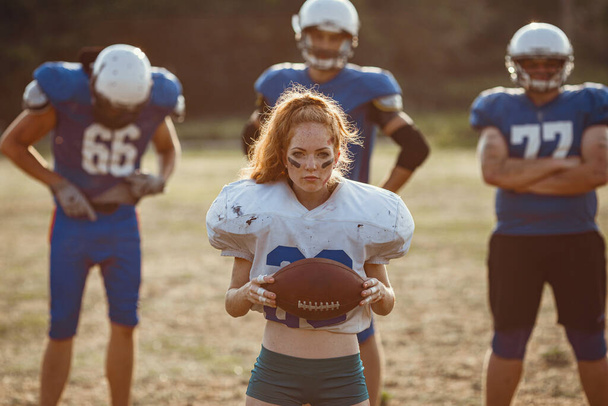 American football woman player in action on the stadium. little red-haired girl with freckles plays sports of large and strong men in football on the field at sunset - Photo, Image