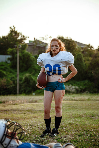 Beautiful young woman wearing American Football attire holding ball. little red-haired girl with freckles plays sports of large and strong men - Photo, image