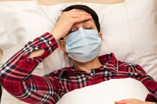Άρρωστη γυναίκα με ιατρική μάσκα ξαπλωμένη στο κρεβάτι που υποφέρει από πυρετό και πονοκέφαλο. Μελαχρινή κοντά μαλλιά με συμπτώματα covid-19 διαμένουν στο σπίτι κατά τη διάρκεια της πανδημίας coronavirus. Σχέδιο καραντίνας. Κλείσιμο - Φωτογραφία, εικόνα