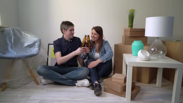 Young couple sitting floor relaxing in apartment drinking beer. Moving new home - Séquence, vidéo