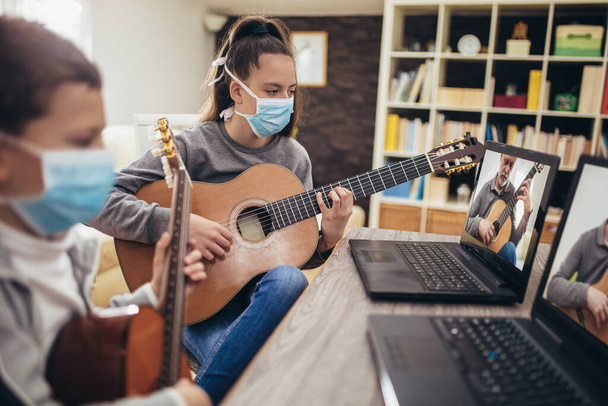 Boy and girl, wear protective masks, playing acoustic guitar and watching online course on laptop while practicing at home. Online training, online classes. - Photo, Image