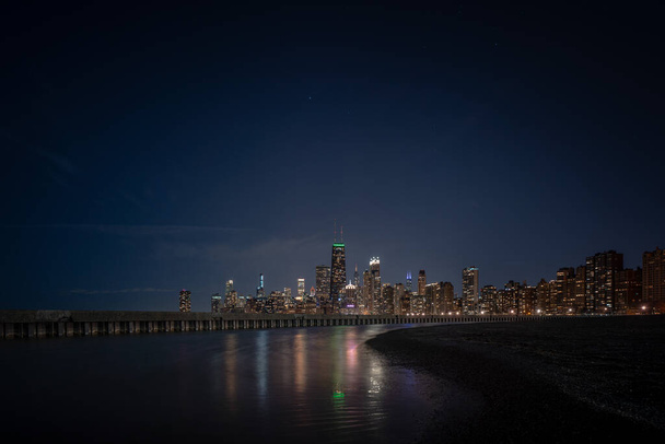 Światła budynków odbijające się od stojącej wody przylegającej do falistej blachy stalowej i betonowego molo z Chicago Skyline na horyzoncie i gwiazdami na ciemnym niebie nad. - Zdjęcie, obraz