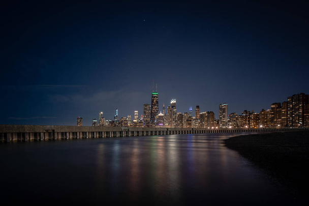 Colorful building lights reflecting off of standing water adjacent to corrugated steel sheet piling and concrete pier with the Chicago Skyline on the horizon and stars in the dark sky above. - Photo, Image