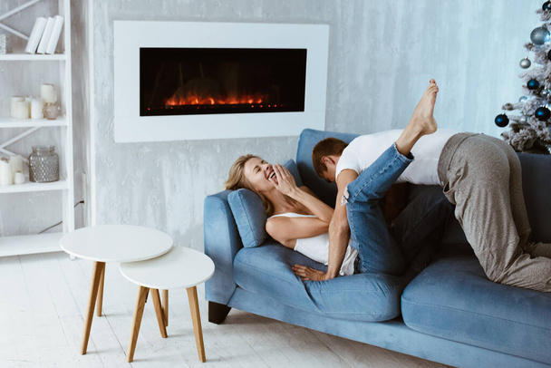 The guy and the girl hug and kiss on the blue couch. Christmas tree decorated with toys. Electronic fireplace. Bright interior. Blonde in a white T-shirt and jeans. - Фото, изображение