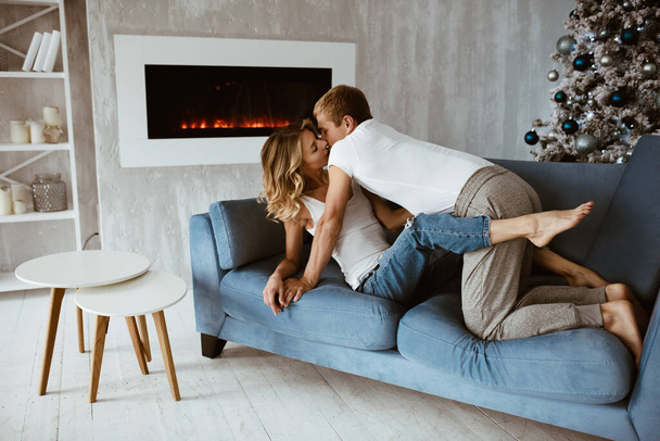 The guy and the girl hug and kiss on the blue couch. Christmas tree decorated with toys. Electronic fireplace. Bright interior. Blonde in a white T-shirt and jeans. - Φωτογραφία, εικόνα