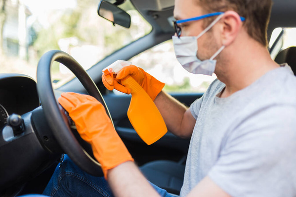 Trabajador con mascarilla facial limpiando volante del coche con paño de microfibra y sosteniendo botella de spray de desinfectante
. - Foto, imagen