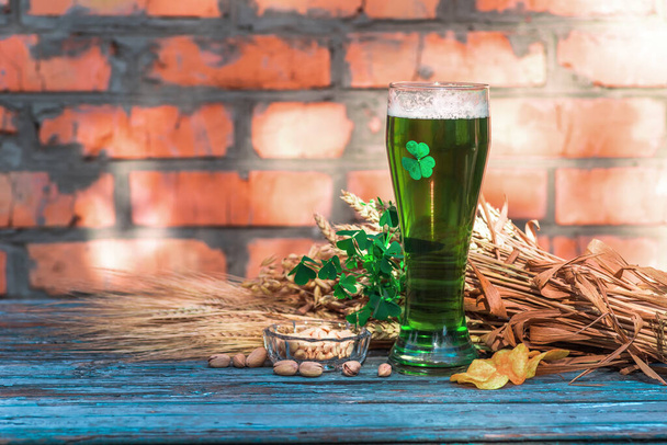 Cerveza verde para el Día de San Patricio contra el fondo de una pared de cerámica se encuentra en una vieja mesa de madera de color junto con ingredientes de la cerveza - trébol, trigo, cebada y bocadillos - pistachos, patatas fritas
 - Foto, imagen