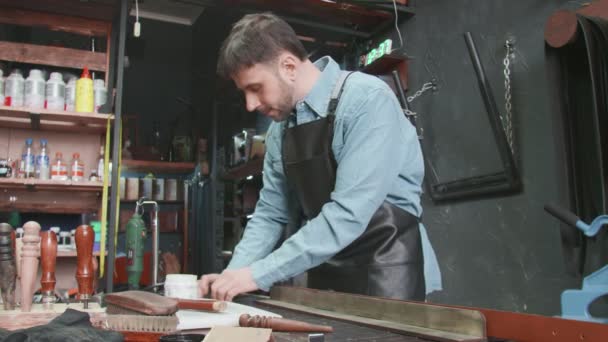 Un tanneur professionnel met de la pâte sur le bord d'une ceinture en atelier. Processus de travail de la ceinture en cuir dans l'atelier de cuir
. - Séquence, vidéo