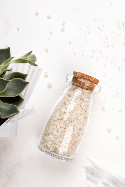 Glass bottle of bath salts. Succulent and leaves on white marble background. Cosmetic spa concept, flat lay or top view - Photo, Image