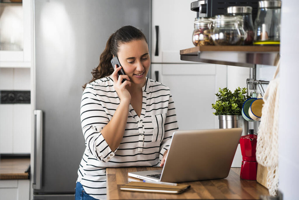 Happy young woman talking on her smartphone and using laptop in her apartment kitchen. Working at home and staying at home. - Photo, image