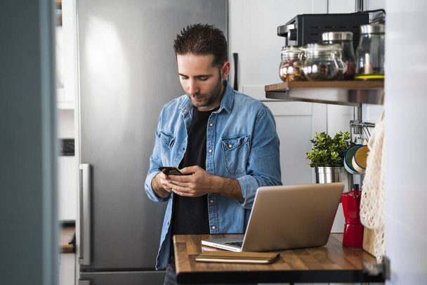 Young man using his smartphone working with laptop in his apartment kitchen. Working at home and stay at home. - Foto, Bild