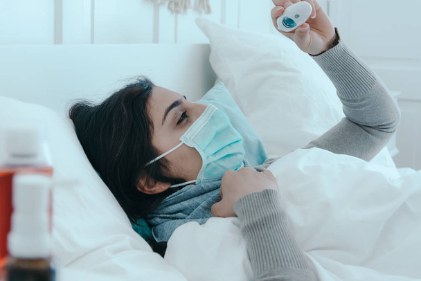 Młoda, chora kobieta leży zmęczona w łóżku w masce i patrzy na termometr kliniczny. . - Zdjęcie, obraz
