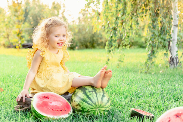 Gelukkig lachend meisje met watermeloenen bij zonsondergang op de boerderij. Gelukkige jeugd. blank kind meisje 3 jaar oud krullend blond rest genieten plezier spelen in geel jurk met watermeloen. - Foto, afbeelding