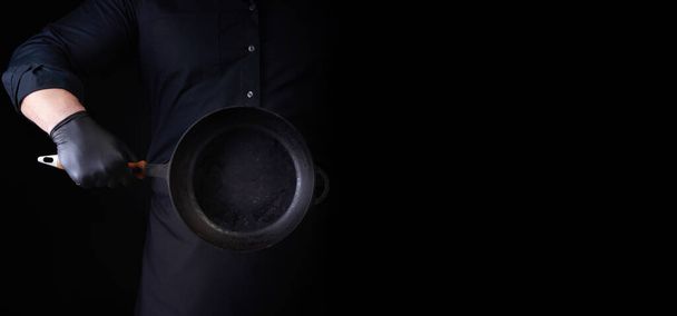 黒の制服とラテックス手袋の男性料理は、彼の前に空のラウンドヴィンテージ黒鋳鉄鍋を保持しています,低キー,そのメニューをラベル付けするレストランのための場所,料理のスケジュールや価格 - 写真・画像