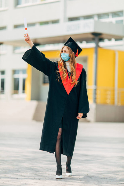 A female graduate student in a protective medical mask, in a black graduation dress, with a diploma in her hands. Graduation ceremony concept, quarantine, coronavirus - 写真・画像