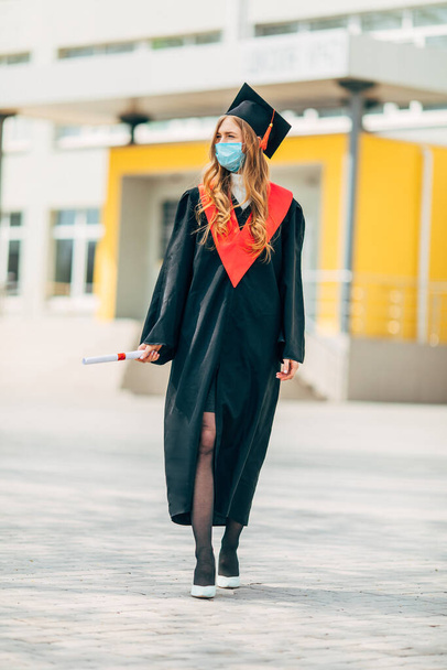 A female graduate student in a protective medical mask, in a black graduation dress, with a diploma in her hands. Graduation ceremony concept, quarantine, coronavirus - Фото, зображення