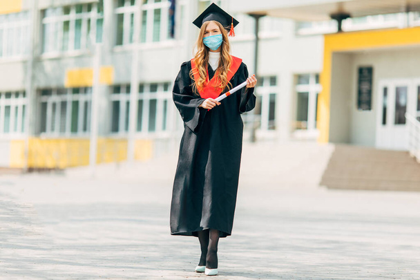 Una estudiante graduada con una máscara médica protectora, con un vestido de graduación negro, con un diploma en sus manos. Concepto de ceremonia de graduación, cuarentena, coronavirus
 - Foto, imagen