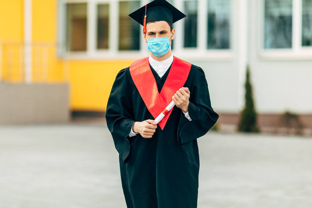 Uczeń-absolwent nosi maskę ochronną przed koronawirusem, w czarnej sukience dyplomowej, z dyplomem w rękach. Pojęcie ceremonii ukończenia szkoły, kwarantanny, koronawirusu - Zdjęcie, obraz