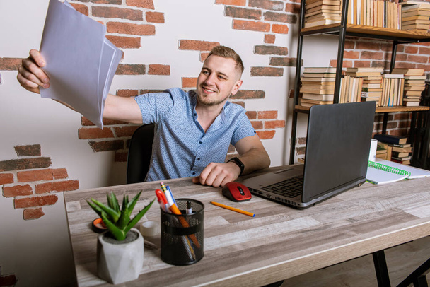Young male businessman working remotely in office, sitting at desk, holding white sheet of paper, cheerful, surprised, smiling - Фото, изображение