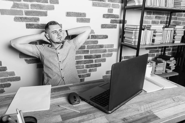 Joven hombre de negocios trabajando remotamente en la oficina, sentado en el escritorio y recostado en la silla y sosteniendo los brazos cruzados detrás de la cabeza, sorprendido, sonriente, cansado después del día de trabajo
 - Foto, imagen