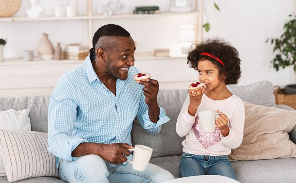 Domowe jedzenie. Afroamerykańskie dziecko jedzące pyszne ciasta z dziadkiem w salonie - Zdjęcie, obraz