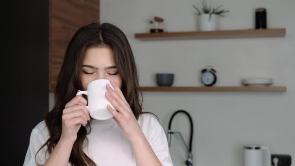 Young woman in kitchen during quarantine. Beautiful positive girl drink from white cup in slow motion. Posing on camera with cheerful emotions. - Filmati, video