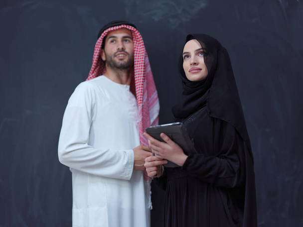young muslim business couple arabian man with woman in fashionable hijab dress using mobile phone and tablet computer in front of black chalkboard representing modern islam fashion technology and ramadan kareem concept - Fotografie, Obrázek