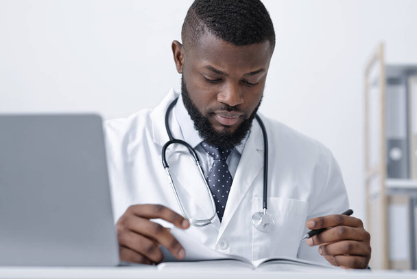 Συγκεντρωμένος μαύρος γιατρός σχεδιάζει την ημέρα του στο γραφείο. - Φωτογραφία, εικόνα