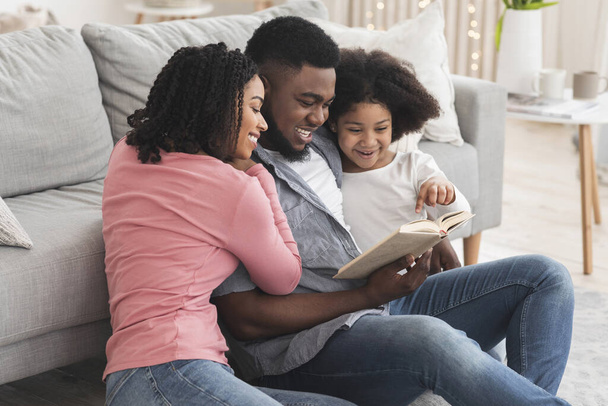 Glückliche schwarze Familie liest zusammen Buch, sitzt auf dem Fußboden im Wohnzimmer - Foto, Bild