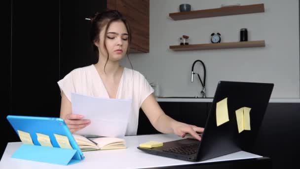Młoda kobieta w kuchni podczas kwarantanny. Poważnie skoncentrowana dziewczyna pisząca na klawiaturze laptopa. Trzymajcie dokumenty w rękach i pracujcie. Praca zdalna w domu. - Materiał filmowy, wideo
