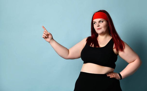 Overweight redhead woman in red headband, black top and leggings. She pointing at something, posing on blue studio background - Foto, Imagem