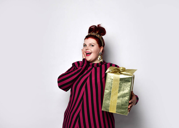 Obese lady in striped dress, crown, earrings. Holding golden gift box tied with ribbon and bow, posing isolated on white - Fotoğraf, Görsel