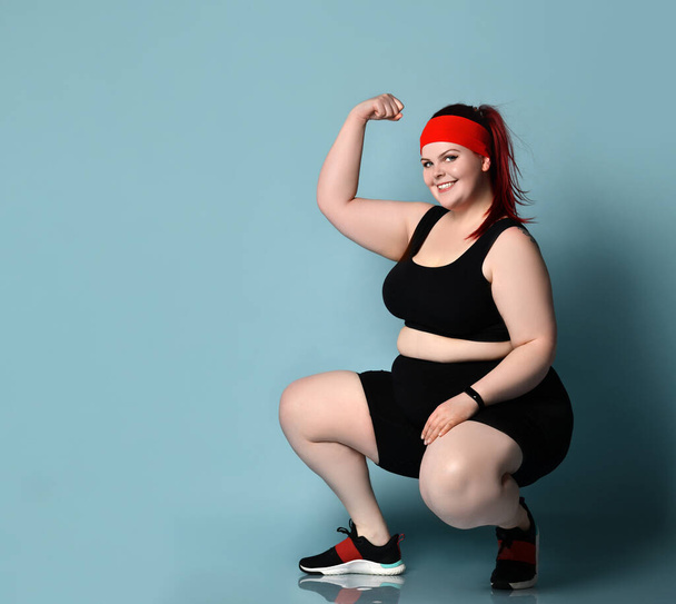 Plus size redhead girl in red headband, black top, shorts, sneakers. Squatting, showing muscles, posing against blue background - Foto, imagen
