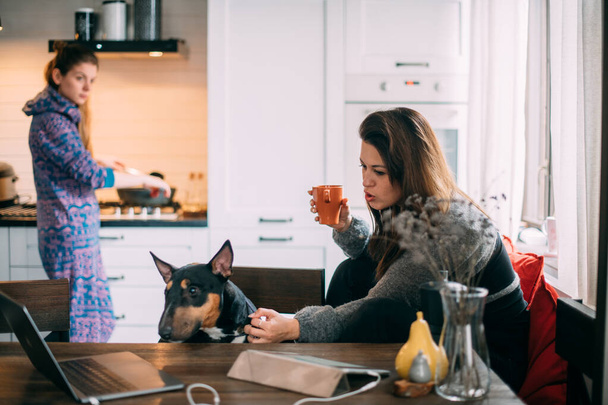 Γυναίκες με σκύλο στην κουζίνα στο σπίτι. Ένα κορίτσι ετοιμάζει φαγητό στο φούρνο, ενώ το άλλο πίνει καφέ και παίζει με το σκυλί στο τραπέζι. - Φωτογραφία, εικόνα