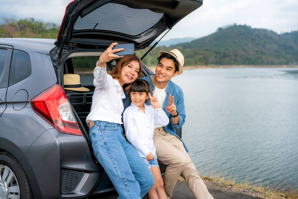 Retrato de la familia asiática sentada en el coche con el padre, la madre y la hija selfie con vista al lago y la montaña por teléfono inteligente, mientras que las vacaciones juntos en vacaciones. Feliz tiempo en familia
. - Foto, Imagen