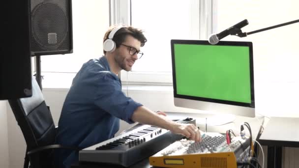 Young man in sound recording studio. Using and adjusting console mixer and enjoying listening to music in headphones. Big green screen. - Footage, Video